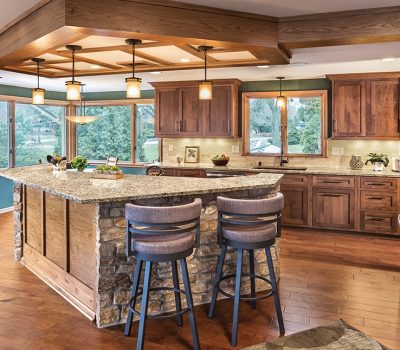 Modern kitchen with wooden cabinetry, stone island, pendant lights, and large windows overlooking a green landscape. Bar stools are neatly arranged.
