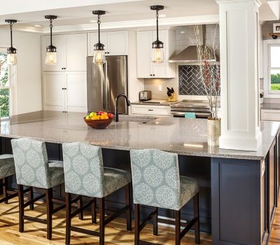 Modern kitchen with large island, pendant lights, and patterned chairs. Stainless steel appliances, white cabinets, and wood flooring create a bright, inviting space.