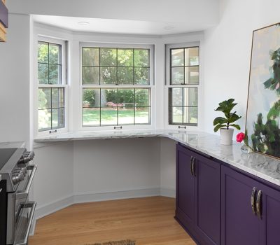 A modern kitchen with marble countertops, purple cabinets, abstract painting, and large bay window view of a green backyard, creating a cozy atmosphere.