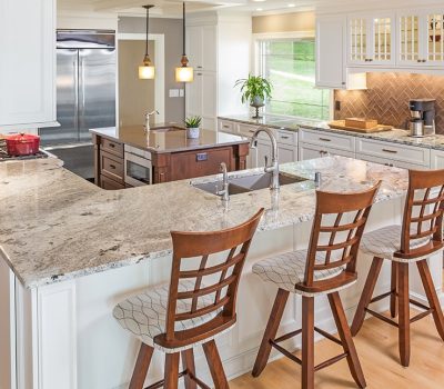 Spacious modern kitchen with granite countertops, wooden bar stools, pendant lighting, large fridge, and greenery outside window. Bright, organized, and inviting space.