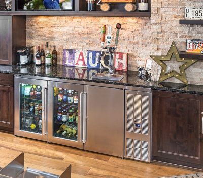 A home bar with dark wood cabinets, stainless steel fridge, bottles, illuminated star, and laugh sign. Decor includes vintage license plates.