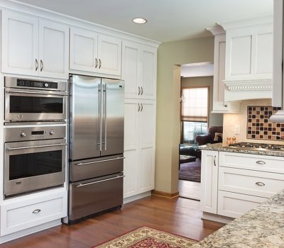 Modern kitchen with white cabinets, stainless steel appliances, and granite countertops. Mixer on counter, living area visible through doorway, wooden floors.