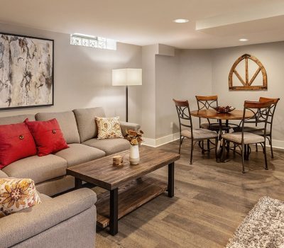 Cozy basement living area features a sectional sofa, colorful cushions, abstract artwork, wooden coffee table, dining set, and warm lighting.