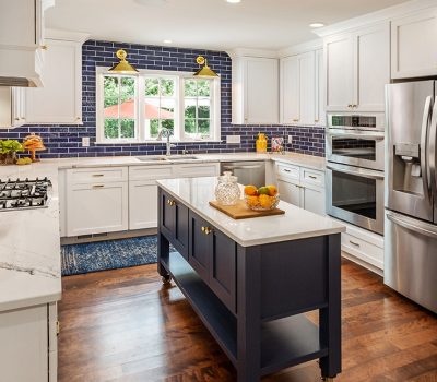 A modern kitchen with white cabinets, blue-tiled backsplash, stainless steel appliances, an island, and large windows overlooking green trees and a red umbrella.