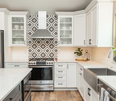 Modern kitchen with white cabinets, geometric backsplash, stainless steel appliances, and farmhouse sink. Bright natural light from window, open shelving with dishes.