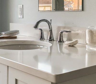 A clean bathroom countertop with a sink, faucet, brush, cotton swabs, and pads in glass containers, reflecting soft natural light.