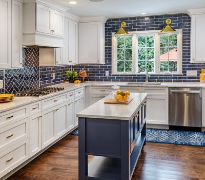 Bright kitchen with white cabinets, blue subway tiles, island with fruit bowl, stainless steel appliances, and large windows overlooking greenery. Warm, inviting atmosphere.