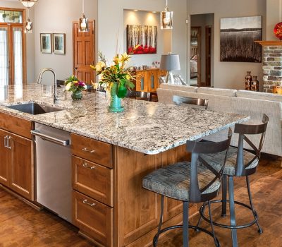 A cozy open-plan kitchen and living area features a granite island, wooden cabinets, sofa, artwork, and a stone fireplace. Elegant and inviting space.