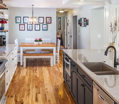 Modern kitchen with granite countertops, white cabinets, and wooden dining table. Artwork decorates walls, and fruit is on the island. Bright and inviting space.