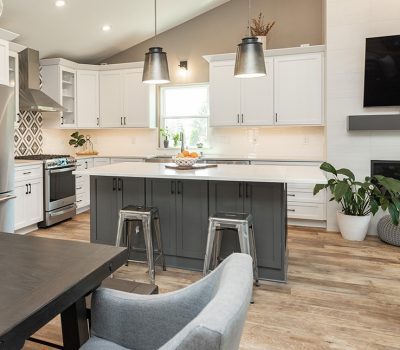 Modern kitchen with white cabinets, stainless steel appliances, and wooden flooring. Features include an island, dining table, plants, and a mounted TV.