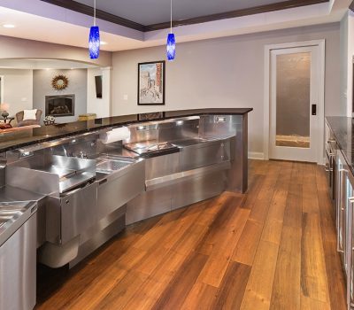 Modern home bar with stainless steel appliances, wooden flooring, blue pendant lights, and a cozy seating area with a fireplace in the background.