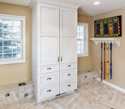 Bright utility room with white cabinets and dog bowls. Colorful animal artwork and leashes on hooks. Windows reveal snowy outdoor view.