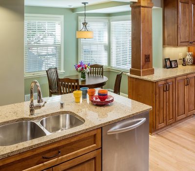 A warm, modern kitchen with wooden cabinets, granite countertops, and a dining table by the window. Colorful dishes and flowers add vibrancy.