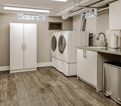 A modern laundry room with a washer, dryer, white cabinets, and a sink. Laminate flooring and natural lighting from high windows.