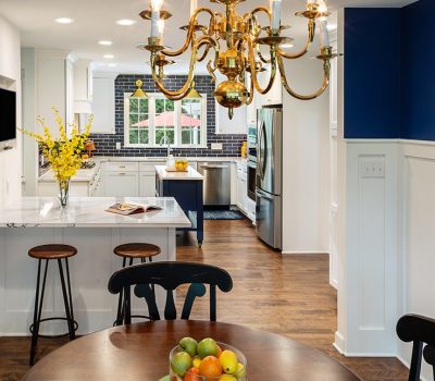 Modern kitchen and dining area with a central chandelier, white cabinetry, dark blue accents, marble countertops, and wooden floors. Vase with yellow flowers.
