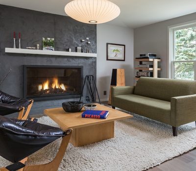 Modern living room with a fireplace, green sofa, leather chairs, and wooden coffee table. Bright window view with trees outside.