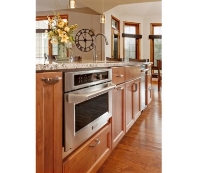 A modern kitchen with granite countertops, stainless steel oven, wooden cabinets, floral arrangement, and decorative wall clock. Bright lighting from windows.