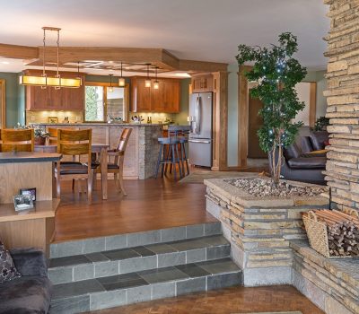 Warm, inviting living space with stone fireplace, wooden accents, and cozy seating. Dining area and modern kitchen visible in open-concept design.