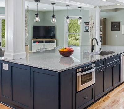 Modern kitchen with large island, gray countertop, and navy cabinets. Open concept design leads to living room with TV and outdoor view.