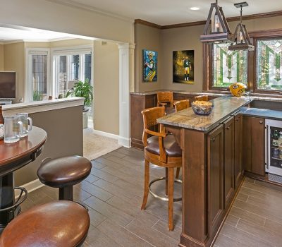 Cozy home bar features wooden furnishings, a small fridge, sports decor, and plants. Adjacent living area with a television and natural lighting.