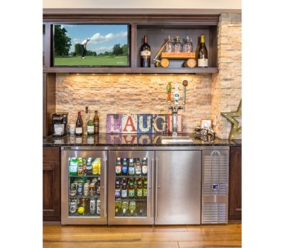Home bar with drink fridges, assorted bottles, and a TV showing golf. Rustic stone backsplash and "LAUGH" decor enhance the setting.