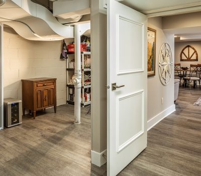 Basement with modern decor, storage shelves, open door leading to a dining area, and decorative wall art. Warm lighting and wooden flooring.