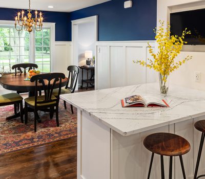 Bright dining room with a round table, floral centerpiece, and blue walls. Adjacent kitchen counter holds a vase and open book.