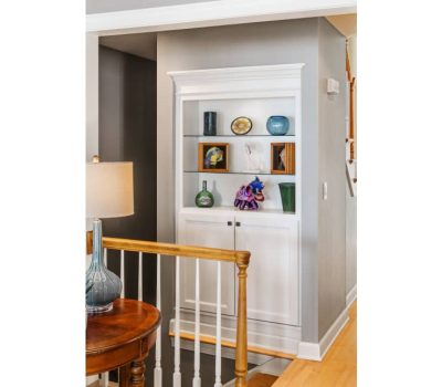 A cozy interior features a white built-in bookshelf displaying colorful vases and art pieces, near a wooden table with a decorative lamp.