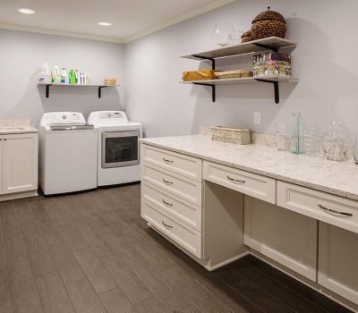 A modern laundry room with a washer, dryer, white cabinets, and countertops. Shelves above hold cleaning supplies and baskets.