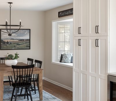 A modern kitchen with wooden table, black chairs, chandelier, landscape painting, white cabinets, large window seat, and decorative plants.