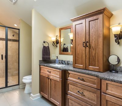 Modern bathroom features wooden cabinets, granite countertops, glass-enclosed shower, and warm lighting. Towel and soap dispenser decorate the double-sink vanity area.