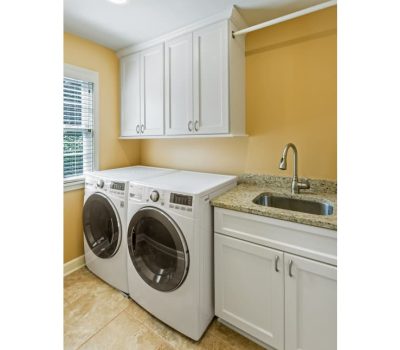 A bright laundry room with orange walls features a washing machine, dryer, white cabinets, a granite counter, and a small sink.