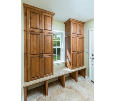 A bright mudroom with wooden cabinets, beige tile flooring, and a window bench under natural light. No people or landmarks are visible.
