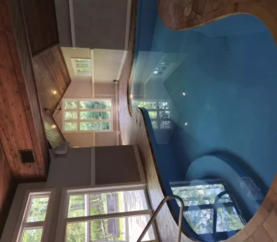 An indoor pool surrounded by wooden walls and large windows. Natural light illuminates the space, with greenery visible outside.