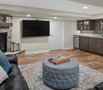 Cozy basement room with a fireplace, a large TV, and a kitchenette. Features a blue ottoman, black couch, and wooden flooring.