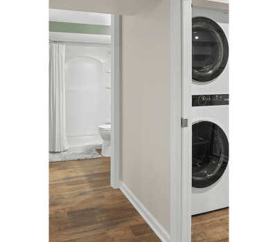 A modern laundry and bathroom featuring a stacked washer-dryer, white shower curtain, toilet, and wooden flooring.
