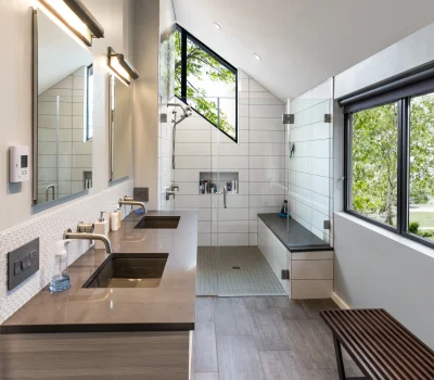 Modern bathroom featuring dual sinks, large mirrors, and a glass-enclosed shower. Natural light enters through angular windows with views of greenery.