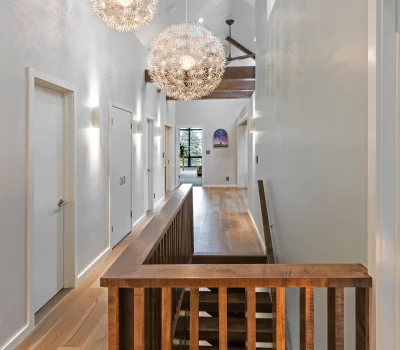 Modern hallway with wooden staircase, stylish spherical chandeliers, and natural light from large windows. Doors line the walls, creating a sleek ambiance.