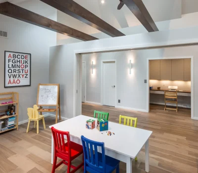 Bright playroom with colorful chairs and table, alphabet chart, easel with drawing, and wooden beams on the ceiling. Cozy, organized space.