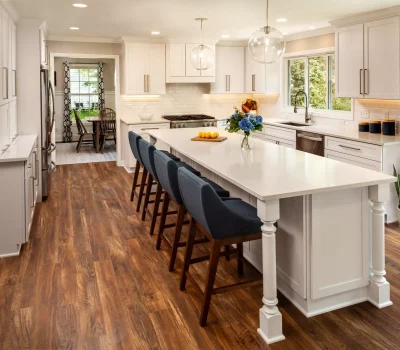 Modern kitchen with wooden flooring, white cabinets, and large island. Four blue chairs, flowers, and lemons add color. Dining area visible through doorway.