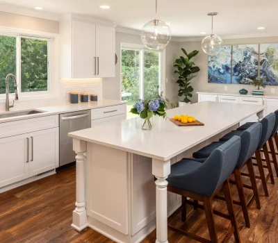 A spacious modern kitchen with white cabinetry, a long island, blue chairs, pendant lights, and a window showcasing greenery outside.