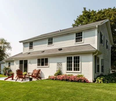 A two-story white house with a well-maintained lawn, patio seating, grill, flowerbeds, and surrounding trees under a clear blue sky.