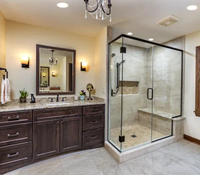 Modern bathroom with glass shower, beige tiles, wooden cabinets, and granite countertop. Decorative plants and towels on the counter. Large wall mirror above sink.