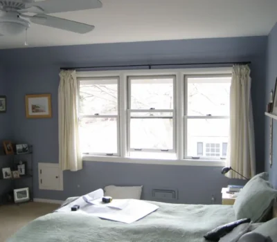 A cozy bedroom with blue walls, a bed, ceiling fan, and large window with light curtains; framed photos and a small shelf present.