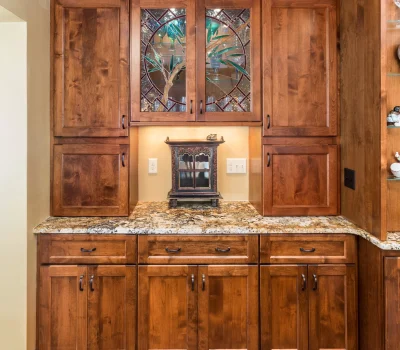 Wooden kitchen cabinets feature intricate stained glass doors and granite countertops, with decorative shelving and hardwood floors complementing the warm interior design.