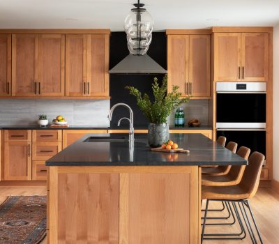 Modern kitchen with wooden cabinets, black countertops, and a central island. Comfortable chairs, stainless steel appliances, and a decorative plant complete the space.