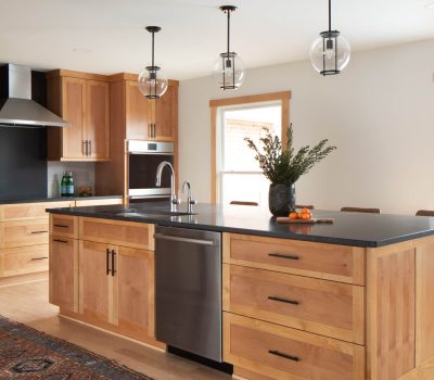 A modern kitchen with wooden cabinets, a black countertop, and stainless steel appliances. Hanging lights illuminate the space, enhancing its warm ambiance.