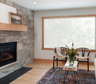 Cozy living room with stone fireplace, wooden mantel, and large window. Vase of flowers on the table, patterned rug on the floor.