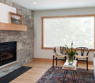 Cozy living room with stone fireplace, framed art, large window, and comfortable seating. Decorative rug and flowers add warmth.