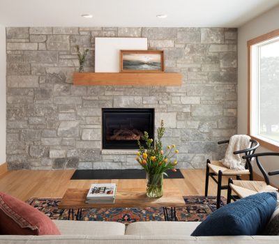 Cozy living room with stone fireplace, wooden mantel, art pieces, and floral arrangement. Natural light from large window illuminates the comfortable seating.